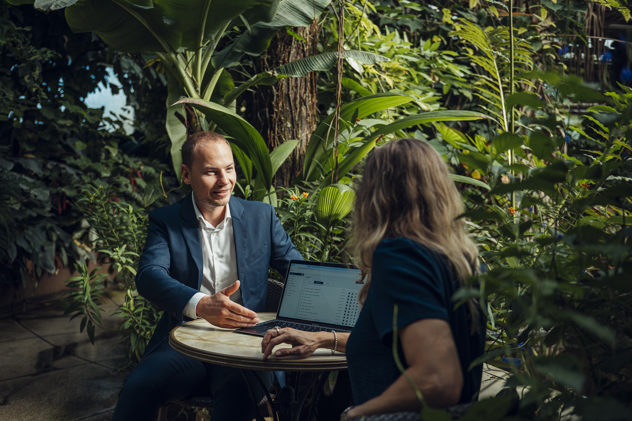Twee professionals in gesprek bij een laptop, omringd door groen: samenwerking en compliance-expertise.